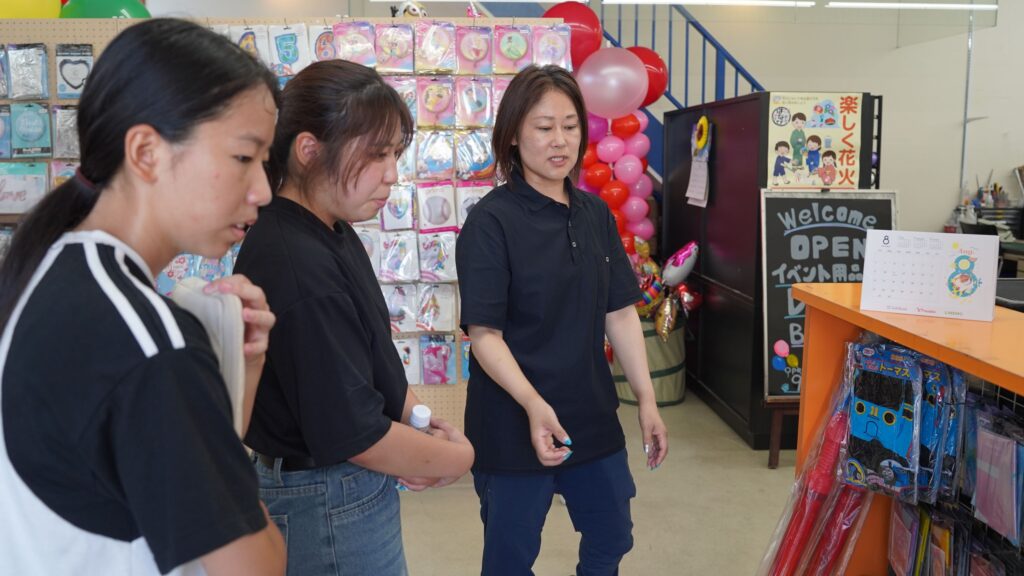 アイデアル株式会社のイベント用品倉庫にて、社員(右・黒)からバルーンやグッズの説明を真剣に受ける学生記者たち(左)。仕事現場を視察。